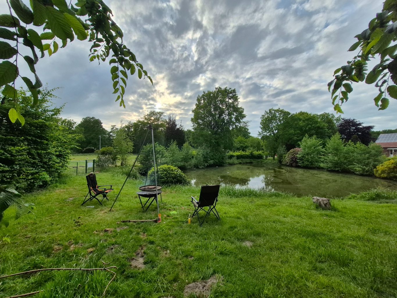 Feuerstelle mit Grill Ruhiger Garten mit See, zwei Stühlen und üppigem Grün unter bewölktem Himmel.
